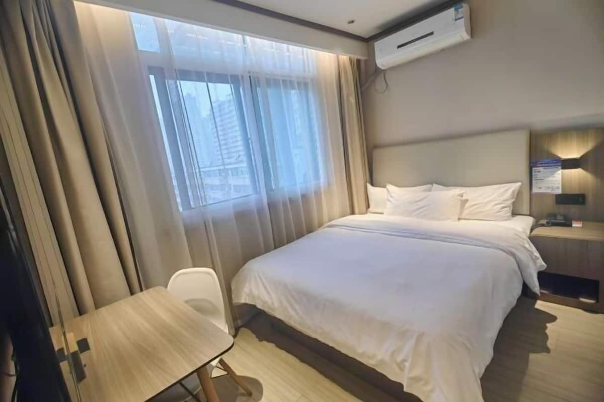 A hotel room with a bed, a desk, a chair, an air conditioner, and a window with curtains.
