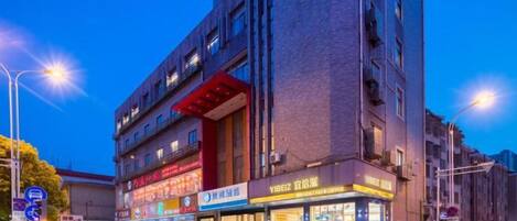 A multi-story building with commercial spaces on the ground floor, including a brightly lit storefront and a parking area with cars.