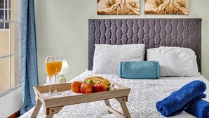 Un letto rifatto con cura, con una testiera grigia, cuscini bianchi e un cuscino decorativo blu. Su un vassoio di legno sono disposti della frutta e bicchieri di succo d'arancia.