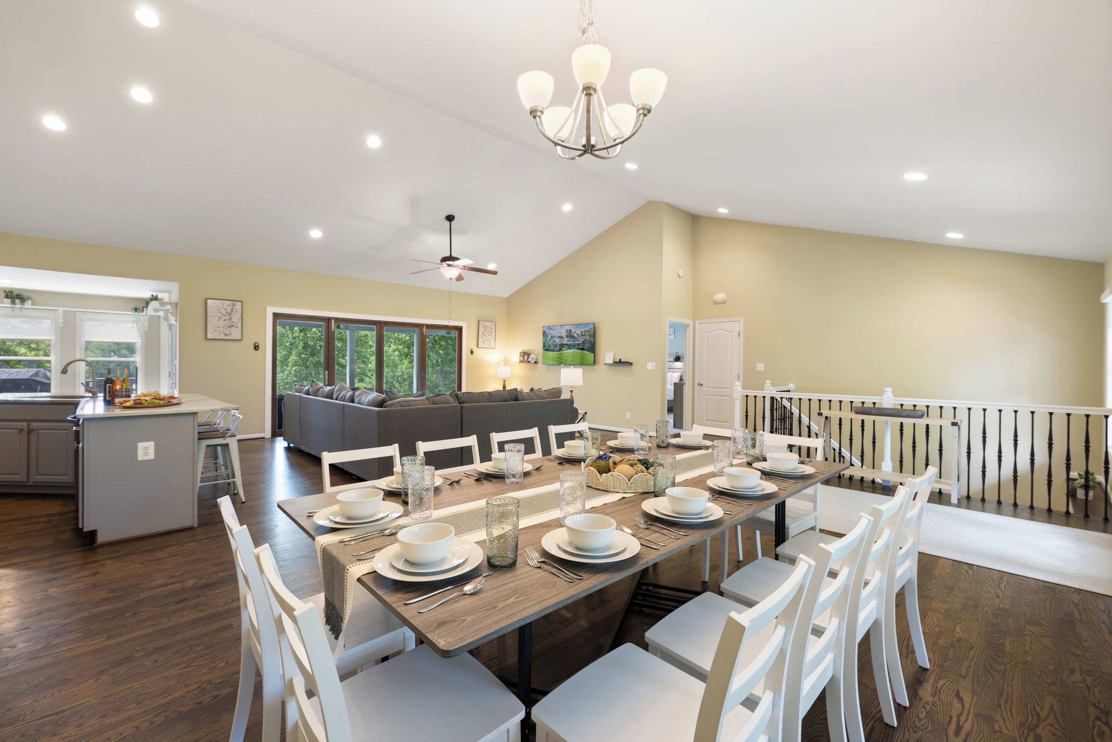 A spacious dining area with a long table set for six, surrounded by white chairs. A chandelier hangs from the ceiling, and a ceiling fan is visible. The room has a high ceiling and large windows allowing natural light.