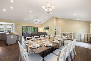 A spacious dining area with a long table set for six, surrounded by white chairs. A chandelier hangs from the ceiling, and a ceiling fan is visible. The room has a high ceiling and large windows allowing natural light.
