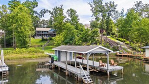 A docked boat with a covered seating area, a small shed, and a wooden pier extending into a calm body of water, surrounded by lush greenery and a residential building in the background. - Lakefront Lake Anna, Dock, Views, Deck, Movies (Mineral)