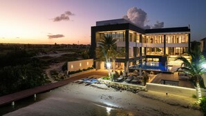 A modern multi-story house with a swimming pool, outdoor seating, and a view of the beach at dusk.