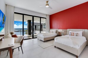 Ein modernes Hotelzimmer mit einem großen Bett, einem kleineren Bett, einem Schreibtisch und einem Sessel. Ein Balkon bietet Blick auf den Ozean.