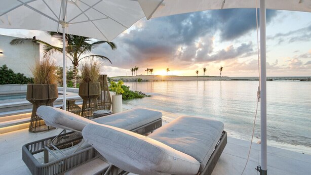 A sunlit outdoor seating area with lounge chairs and umbrellas overlooking a calm body of water and a sunset.