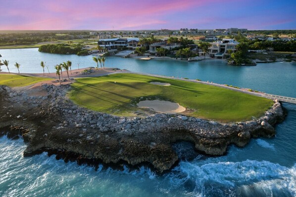 A coastal golf course with a rocky shoreline, a water body, and a residential area in the background.