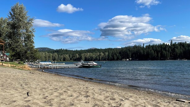 A sandy beach with boats docked near the shore, a forested hill in the background, and a clear blue sky.