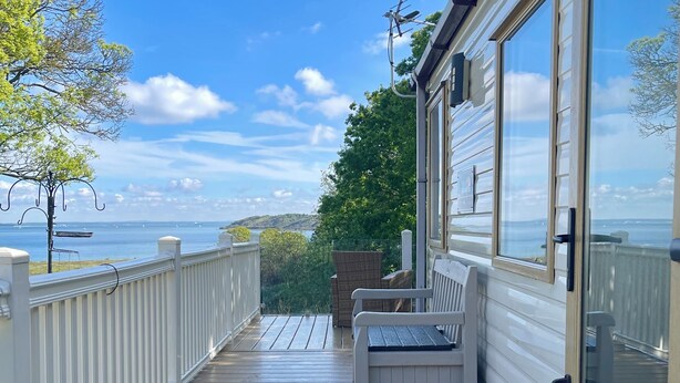 A wooden deck with a white railing, a bench, and a door leading to a glass room. - Paws a While! caravan with uninterrupted views of Solent and direct beach access (Great Thorness)