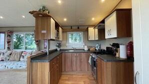 A compact kitchen with wooden cabinets, a central island, and a window with blinds. - Paws a While! caravan with uninterrupted views of Solent and direct beach access (Great Thorness)