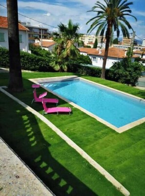 A rectangular swimming pool with a clear blue water surface, surrounded by a well-maintained green lawn. There are two pink lounge chairs positioned on the lawn, facing the pool.