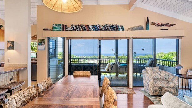 A spacious room with a large wooden dining table, comfortable seating, a bookshelf, and a view of the ocean through large sliding doors. - 180 Degree Ocean View in Wailea! WE204 (Kihei)