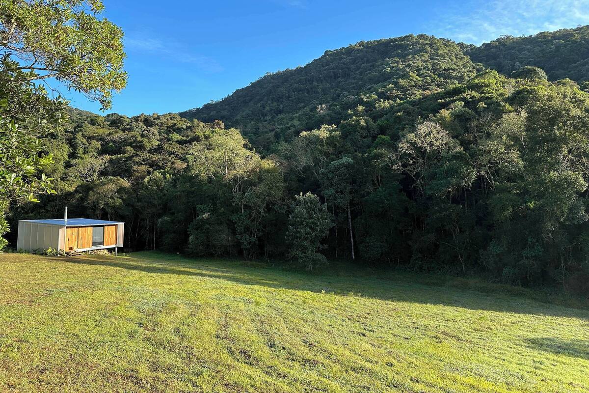A small wooden cabin with a solar panel roof in a grassy field, surrounded by dense forest and hills.