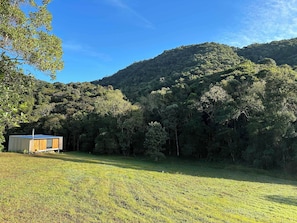 A small wooden cabin with a solar panel roof in a grassy field, surrounded by dense forest and hills. - Clareira Cabanas (Gonçalves)