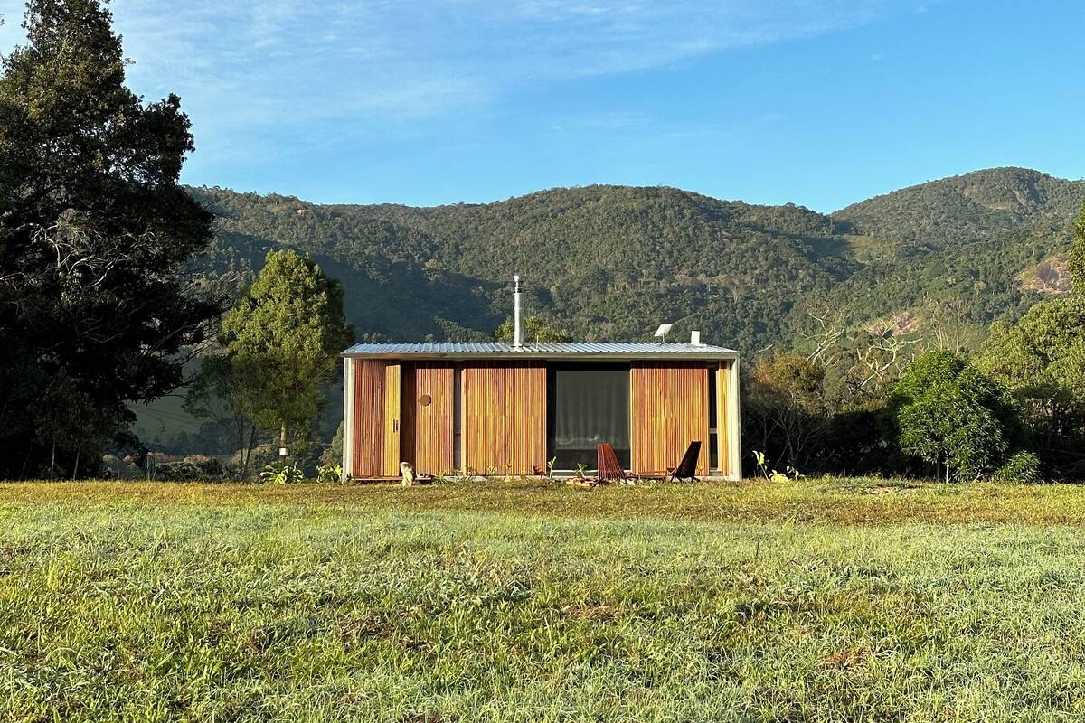 A wooden cabin with a large sliding door, situated in a grassy field with a backdrop of hills and trees.