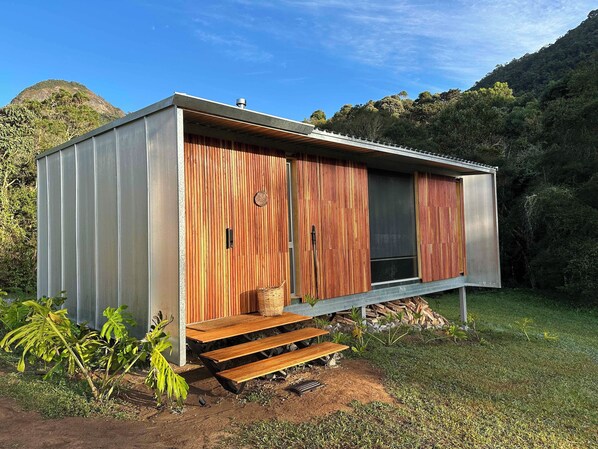 A modern, single-story cabin with a wooden exterior and a metal roof, situated in a grassy area with a forested hill in the background. - Clareira Cabanas (Gonçalves)