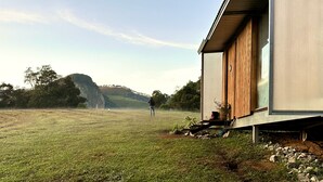 Ein modernes, eingeschossiges Gebäude mit flachem Dach und großen Fenstern, gelegen in einer grünen Wiese vor einer hügeligen Landschaft.