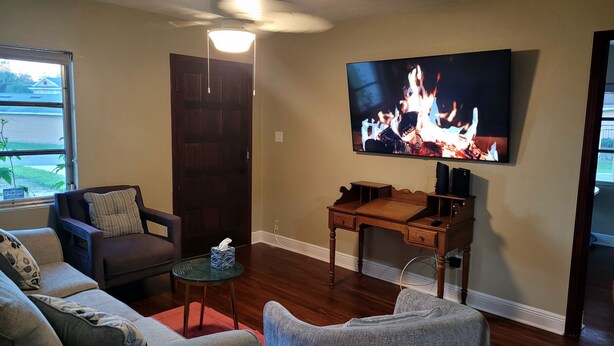 A living room with a TV displaying a fire, a wooden desk, a couch, and a small table. - Cozy Home Near I-4, Lakeland hospital, cancer center ＆ Joker Marchant Stadium (Lakeland)