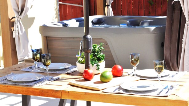 Mesa para dos con platos, cubiertos y vasos, un jacuzzi al fondo y una planta en maceta sobre la mesa.