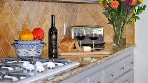 Una encimera de cocina con un tostador, una botella de vino, una hogaza de pan y un bol de fruta.