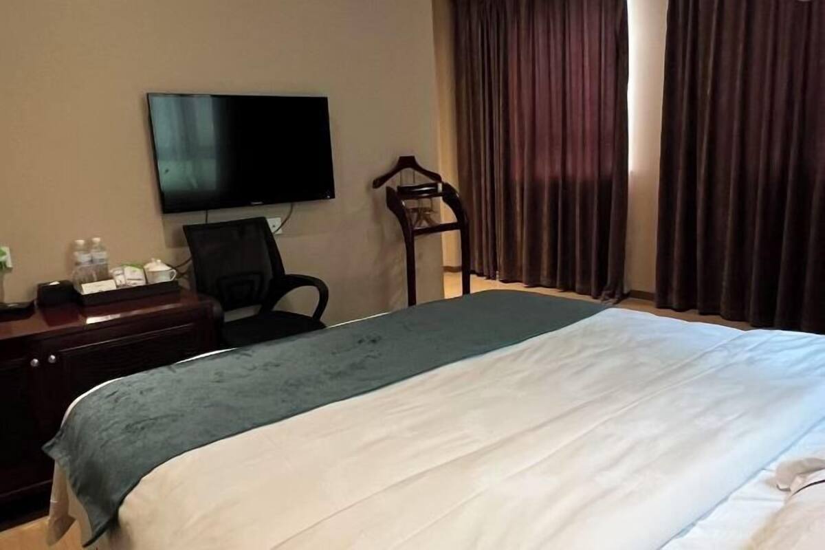 A hotel room with a bed, a television mounted on the wall, a desk with a chair, and a window with curtains.