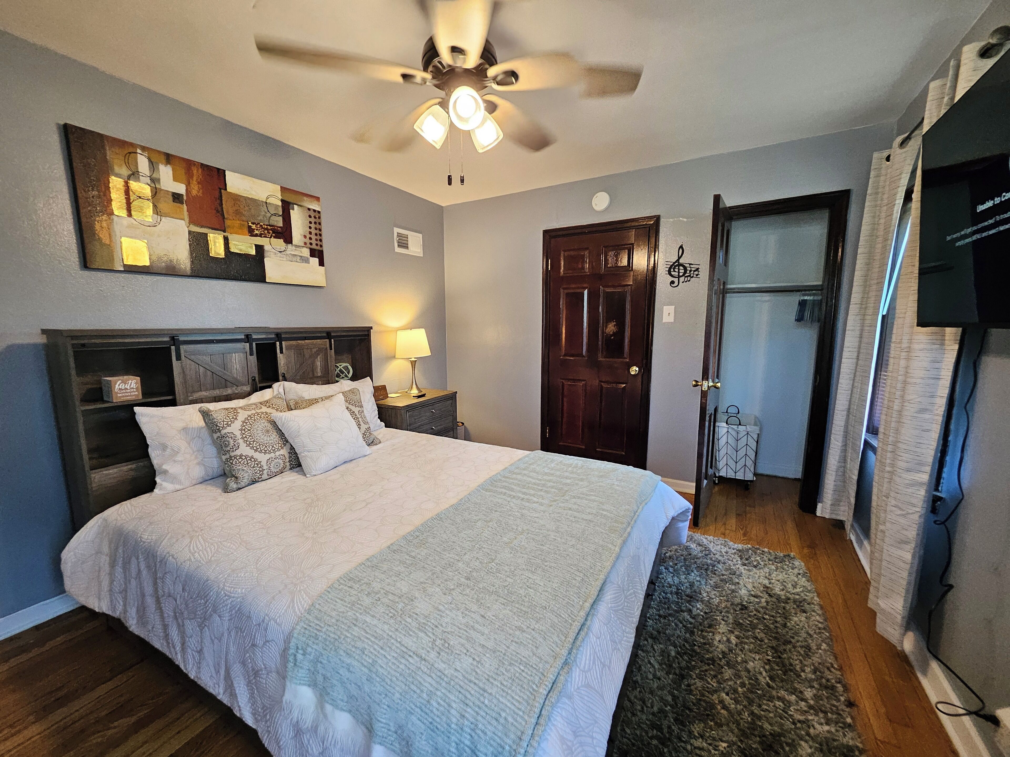 A bedroom with a bed, a ceiling fan, a wooden headboard, a nightstand with a lamp, and a door with a music note decoration.
