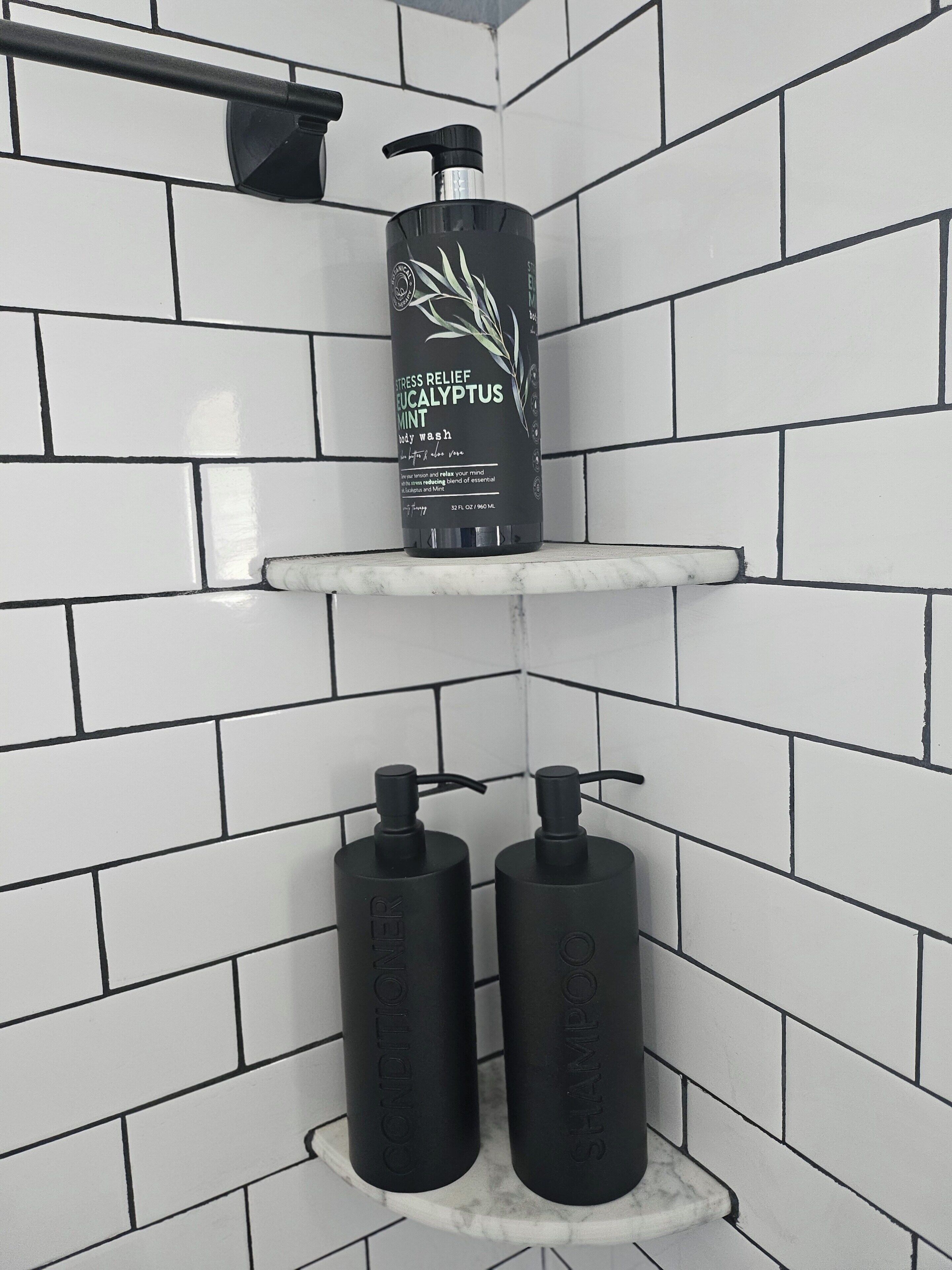 Un baño con paredes de azulejos blancos y un estante de mármol que sostiene una botella de gel de ducha de eucalipto y menta, junto con dos botellas de acondicionador.