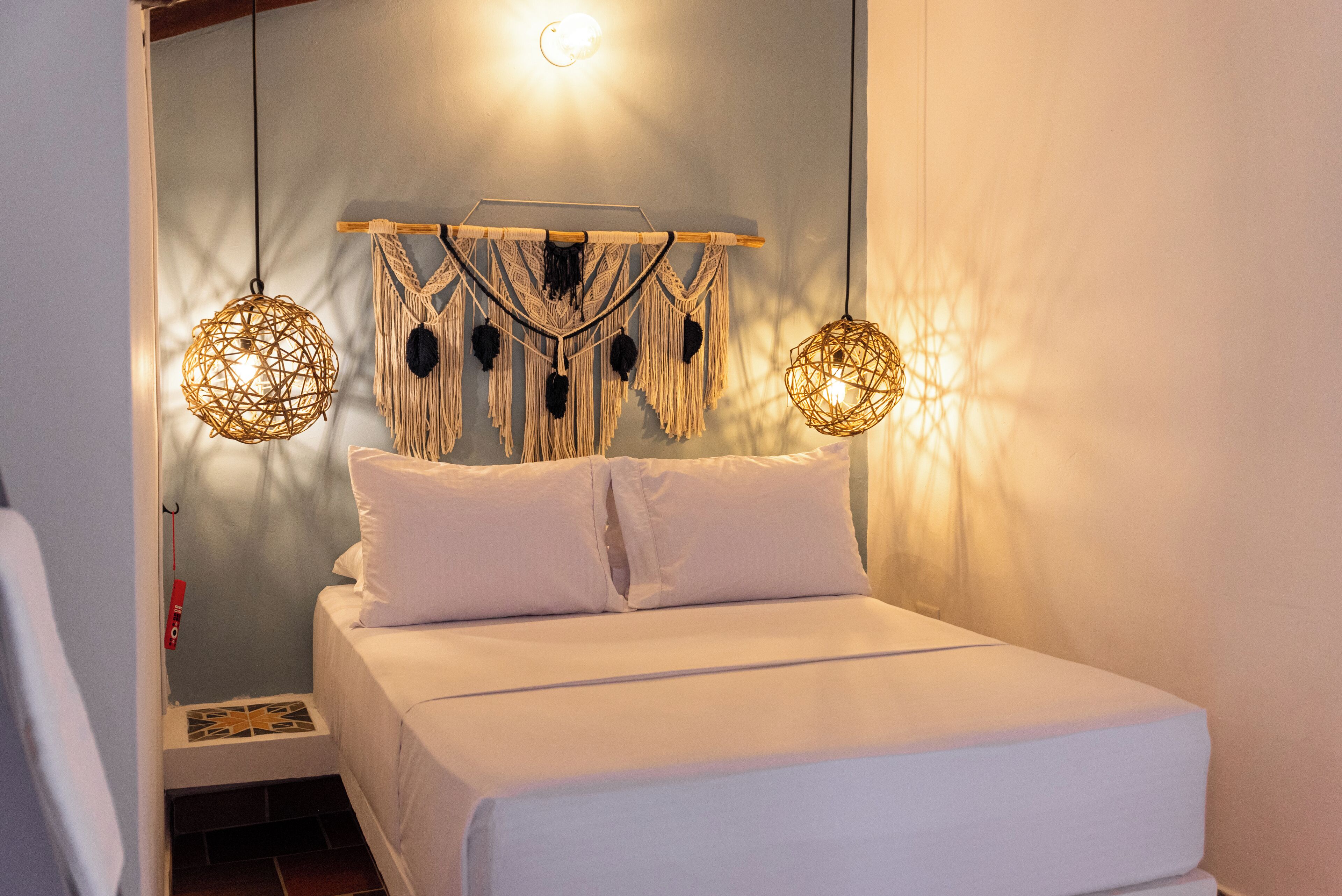 A neatly made bed with white linens, a macrame wall hanging above it, and two woven pendant lights.