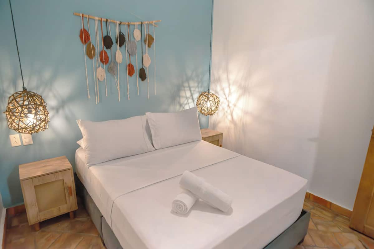 A neatly made bed with white linens, a small wooden nightstand, and a wall hanging with colorful pom-poms.