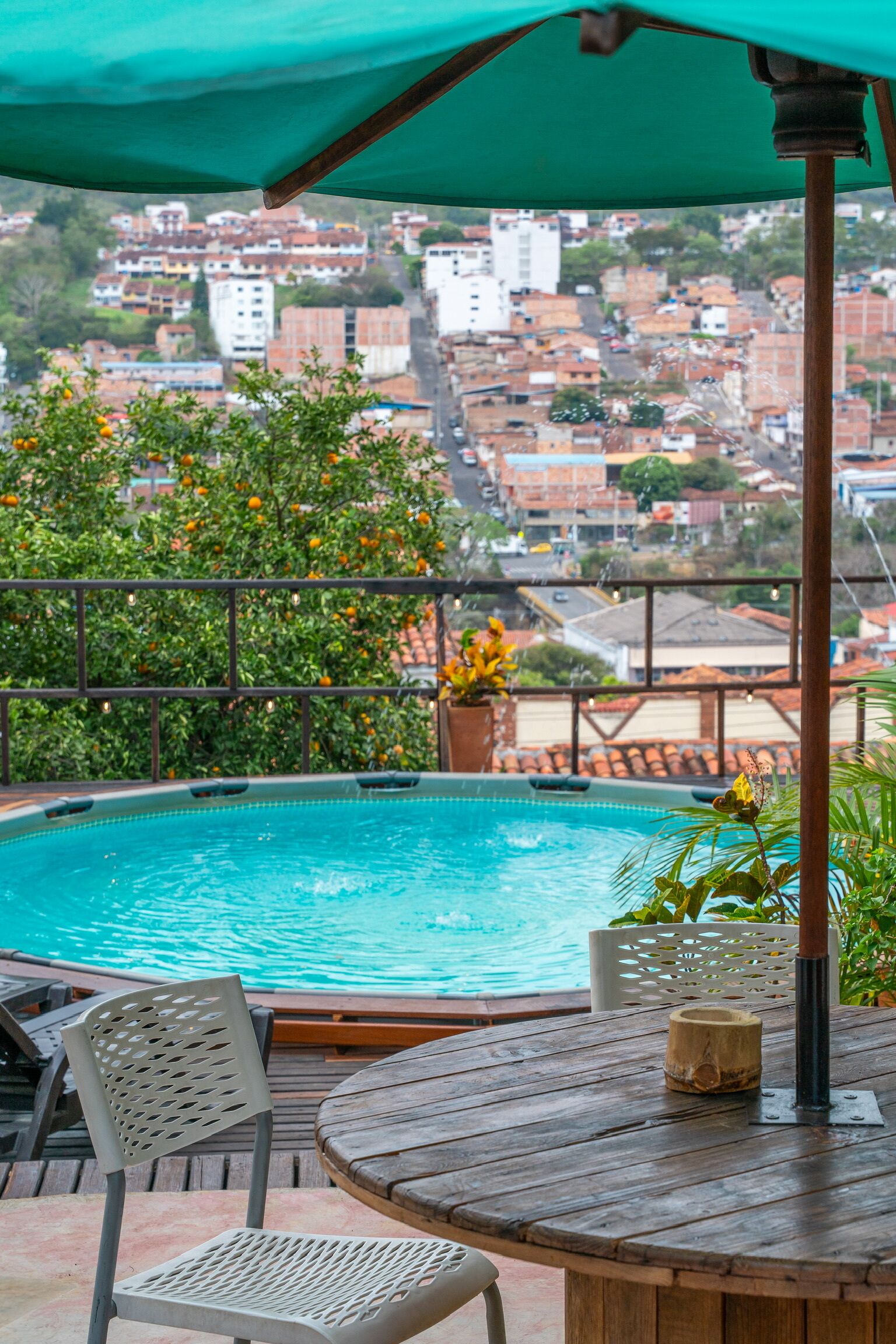 Un área junto a la piscina con una mesa y sillas de madera, una sombrilla verde y vista a un barrio residencial al fondo.