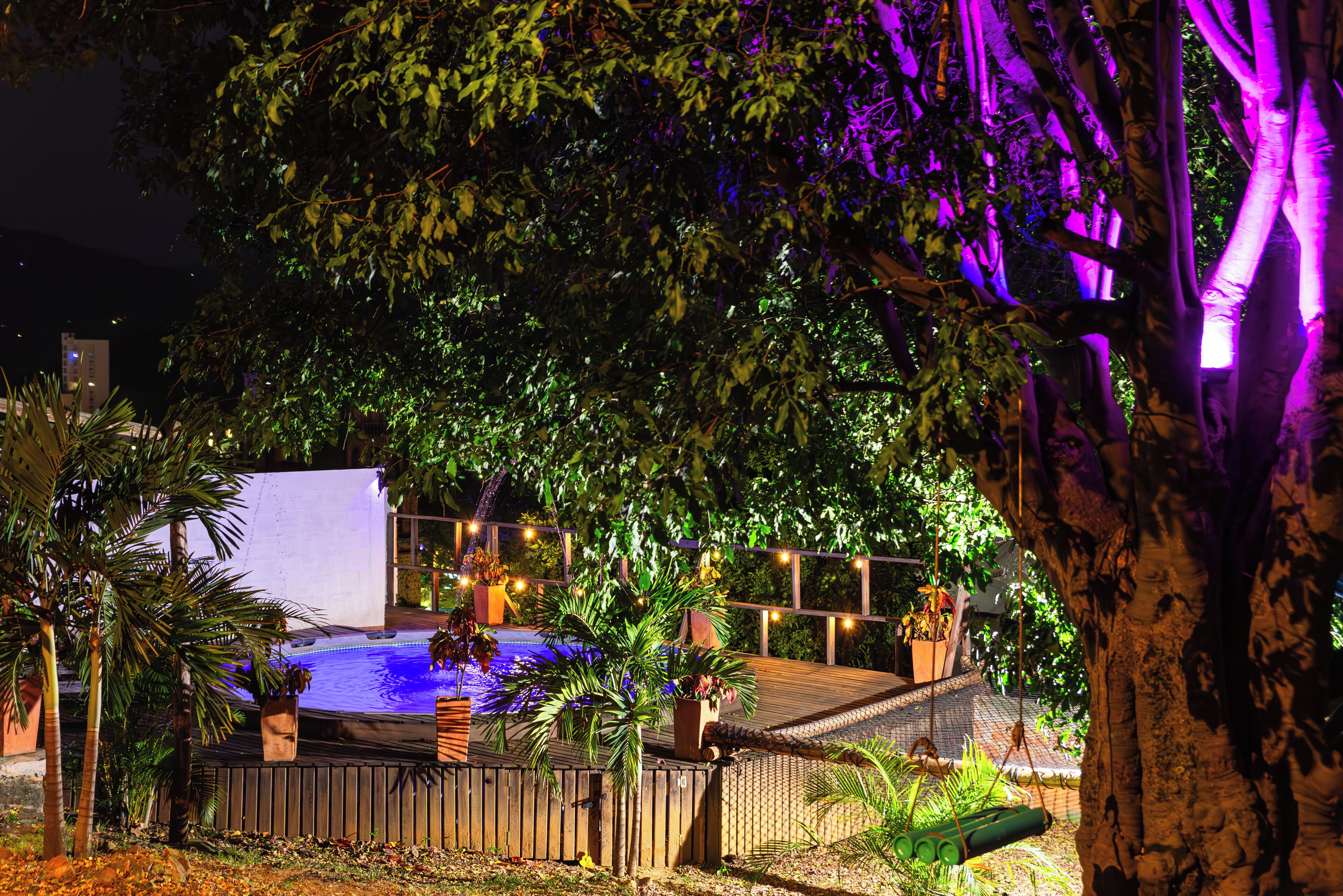 Una piscina circondata da alberi e piante, con un solarium in legno e luci decorative.