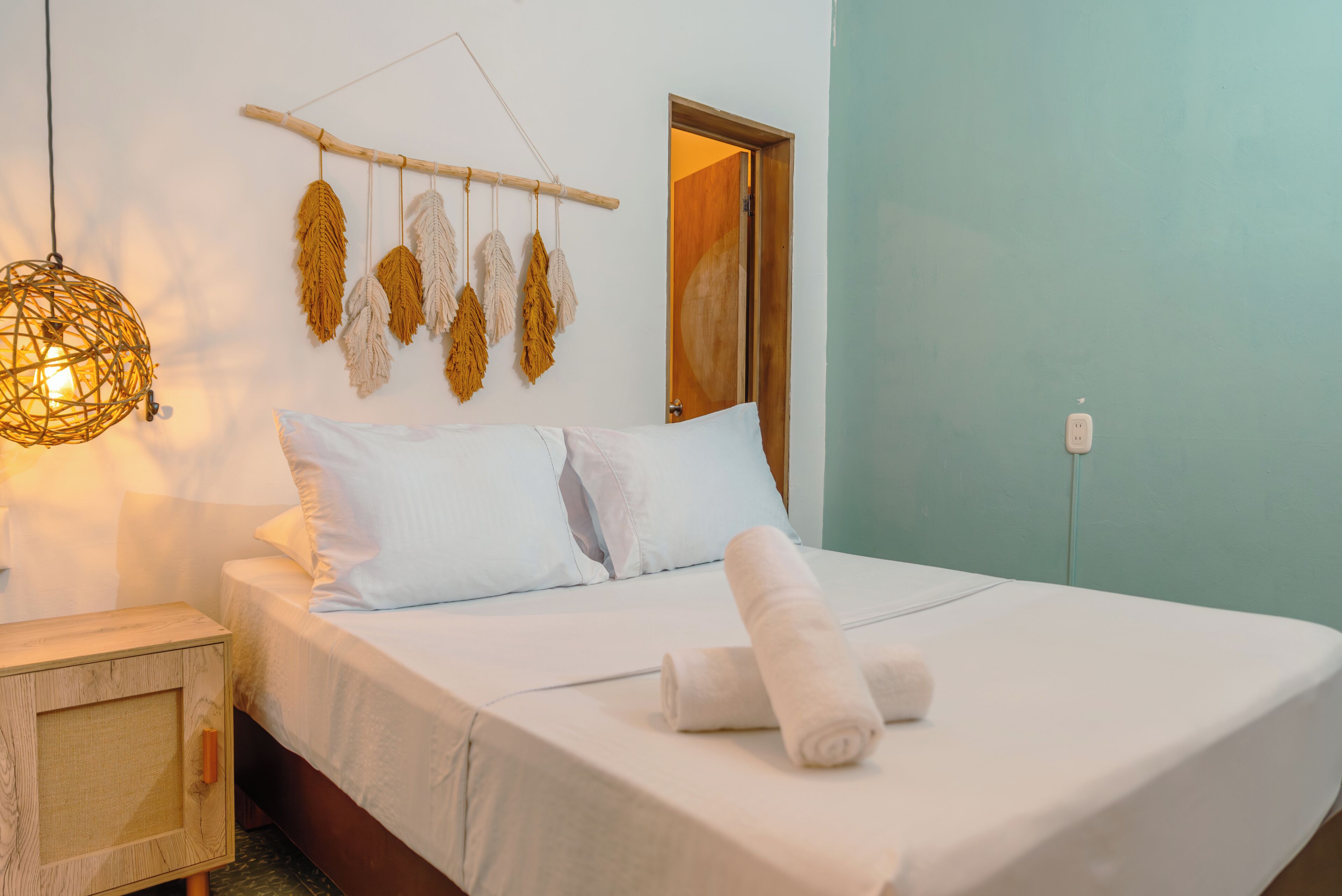 Una cama bien hecha con ropa blanca y una toalla enrollada, una mesita de noche de madera con una lámpara de fibras naturales, y un adorno de macramé en la pared.