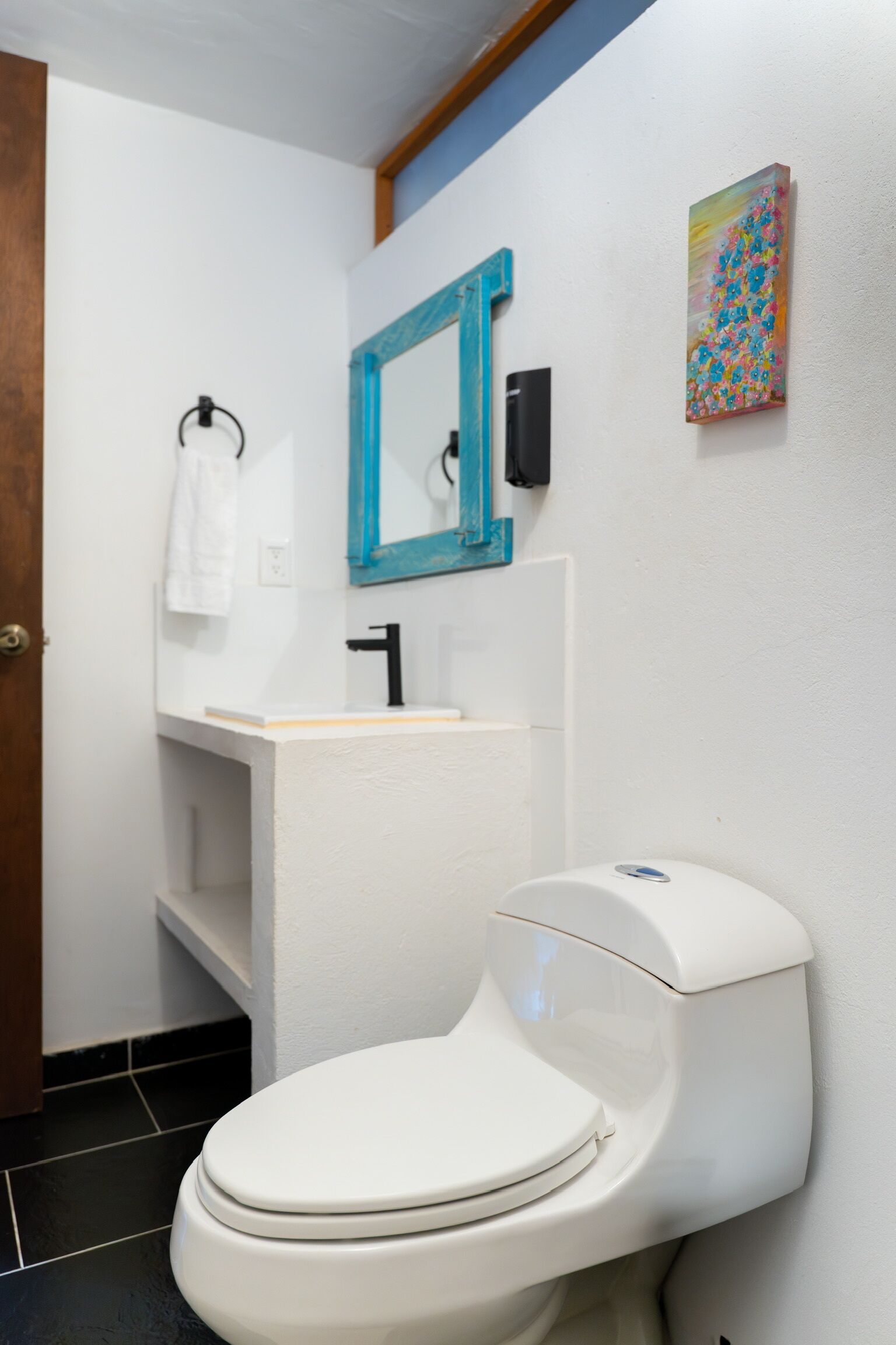 A bathroom with a white toilet, a sink with a blue mirror, and a colorful painting on the wall.
