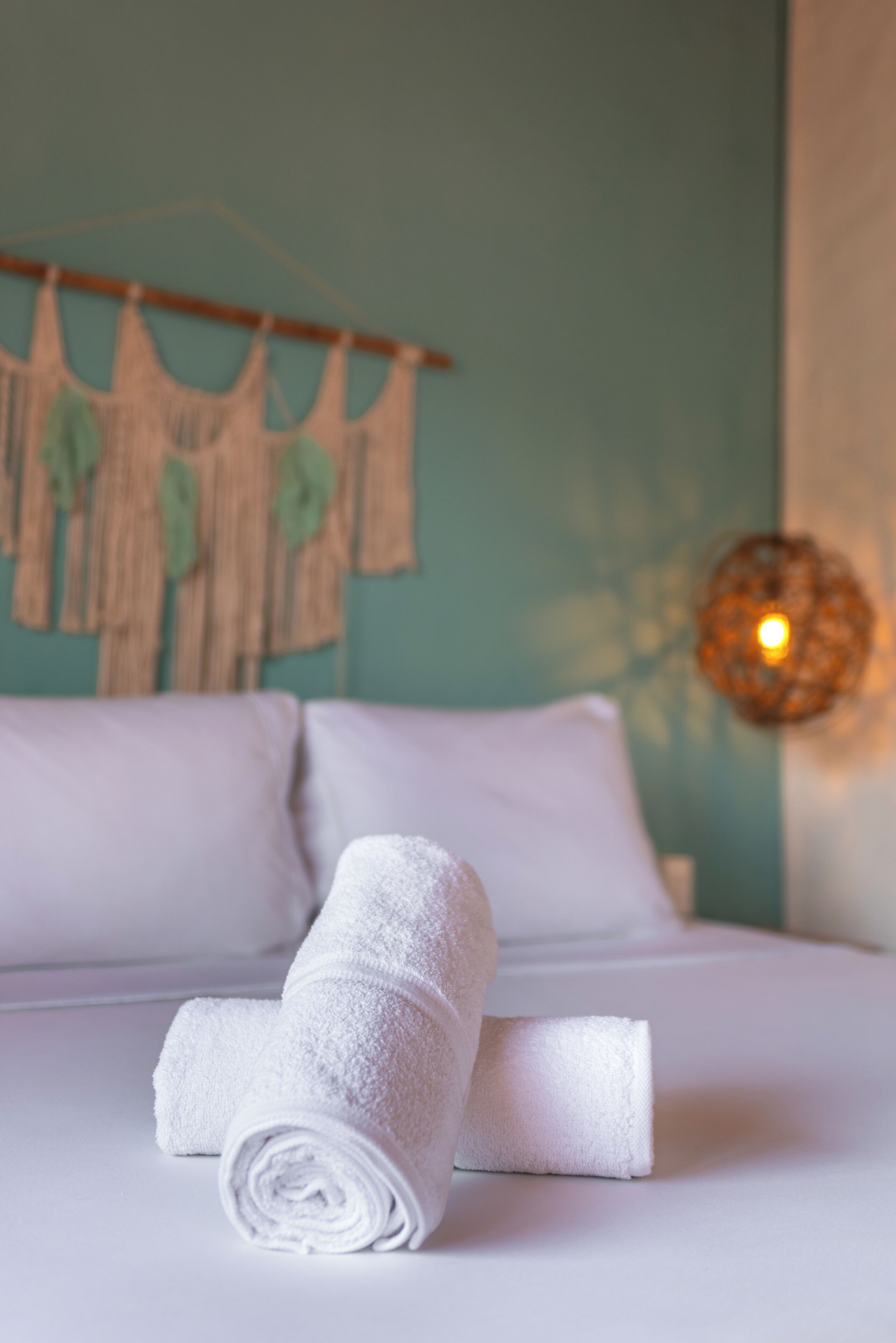 A neatly made bed with white linens, a wall with a macrame piece, and a hanging light fixture.
