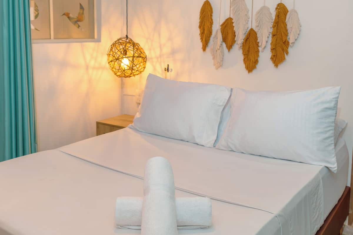 A neatly made bed with white linens and rolled towels, a hanging light fixture, and wall-mounted feather decorations.