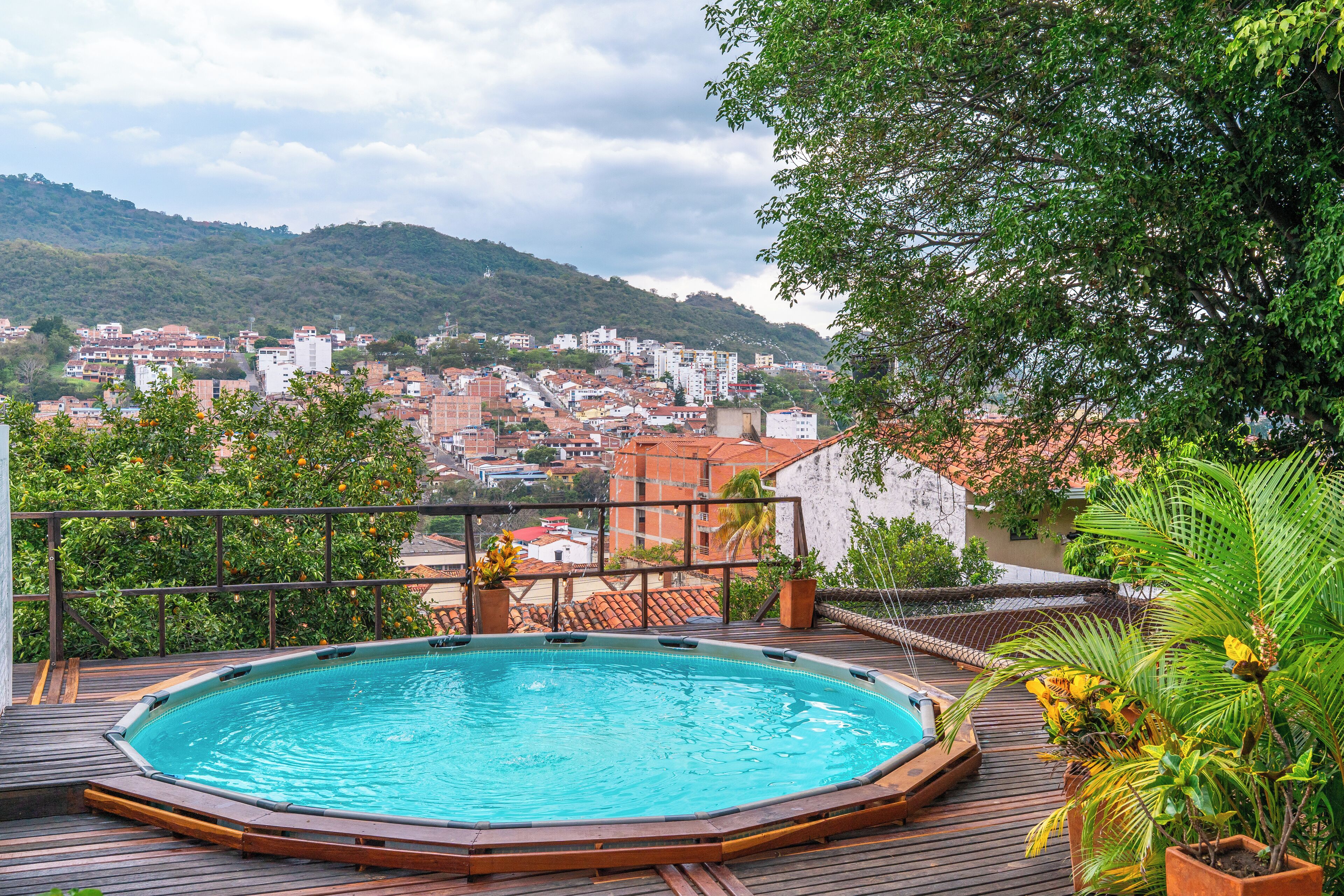 A rooftop swimming pool with a wooden deck, surrounded by potted plants and overlooking a cityscape with buildings and hills.