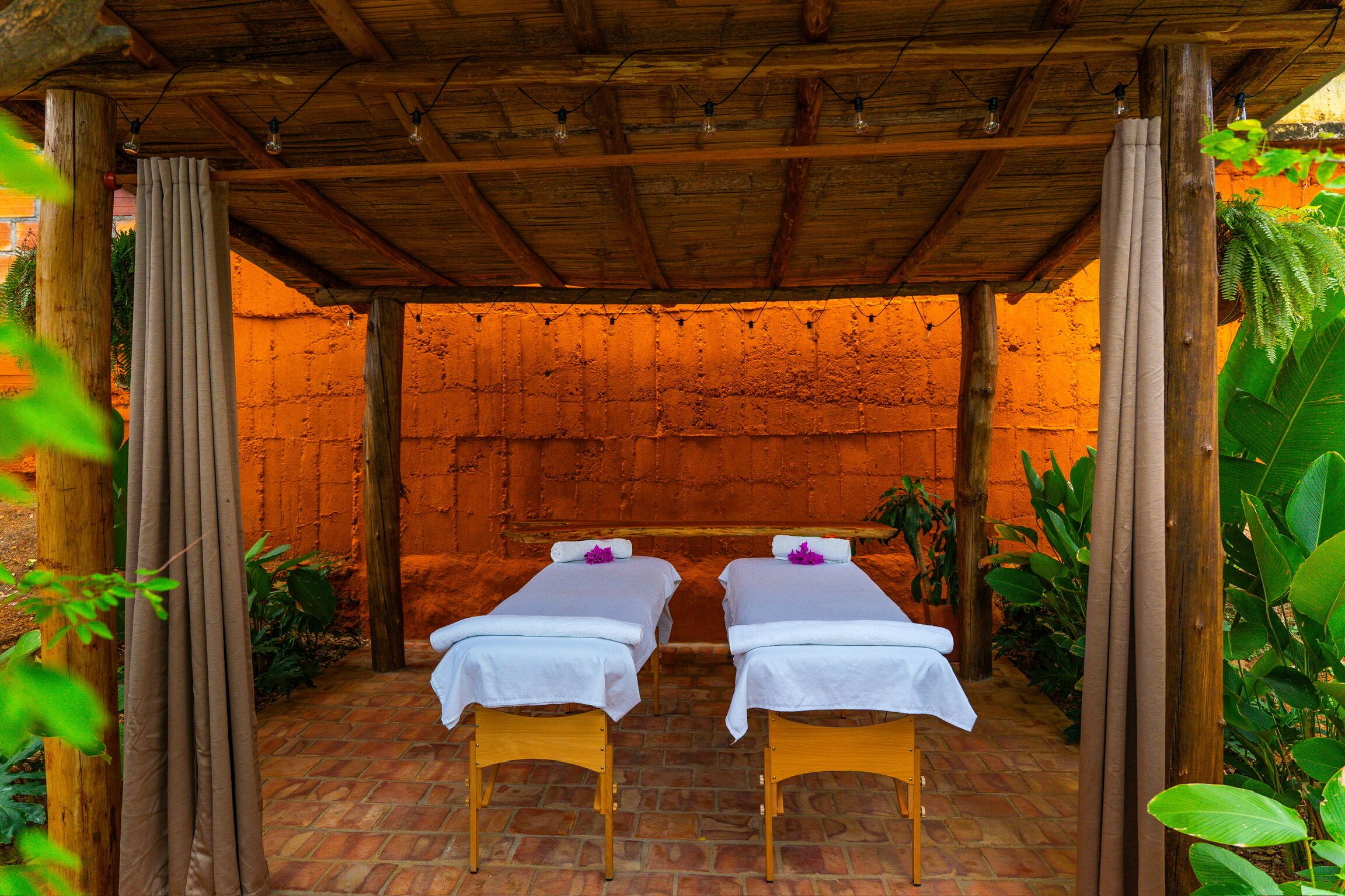 A wooden pergola with two massage tables under a thatched roof, surrounded by lush greenery.