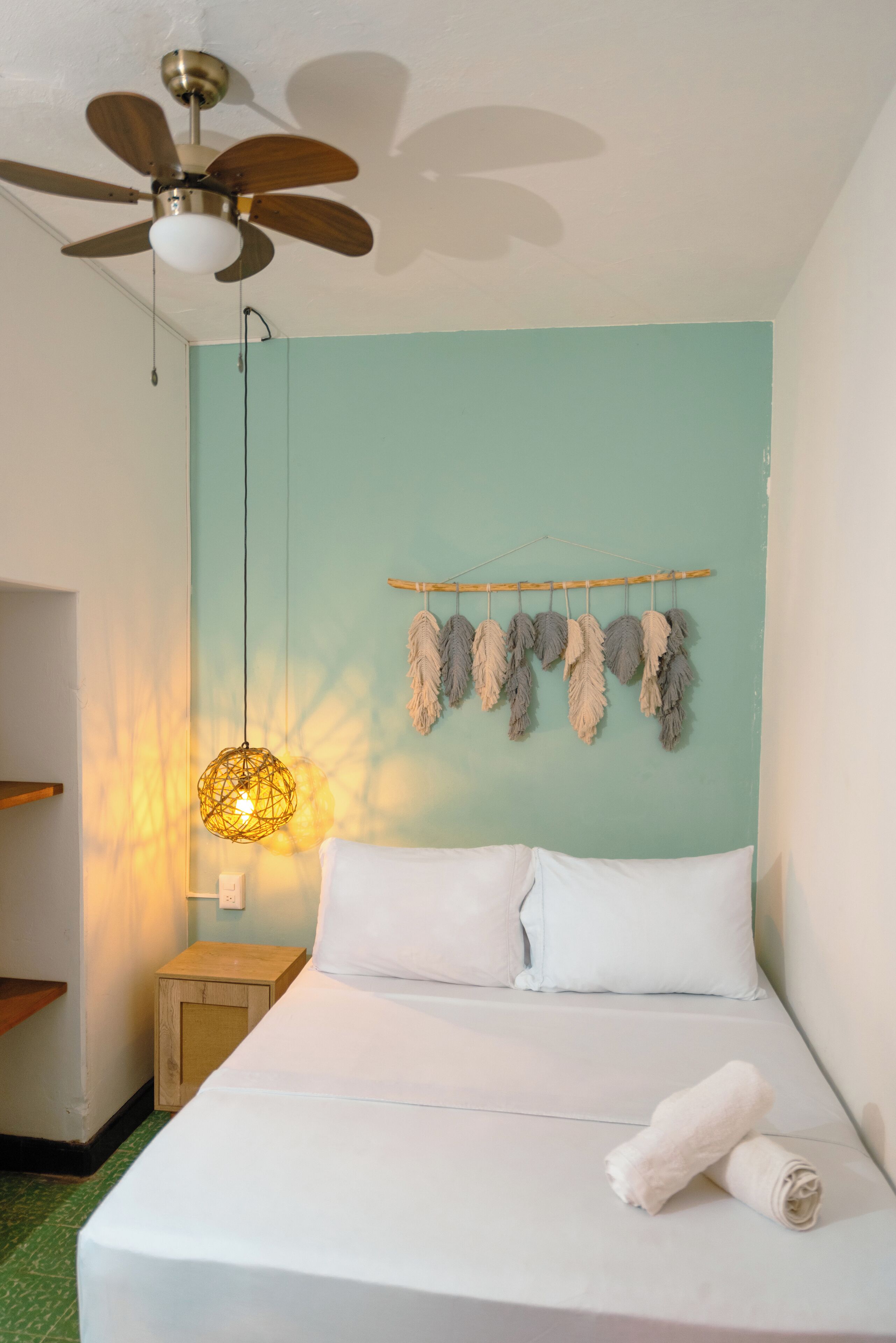 Una cama bien tendida con sábanas blancas, una mesita de noche de madera, una lámpara colgante y un ventilador de techo.