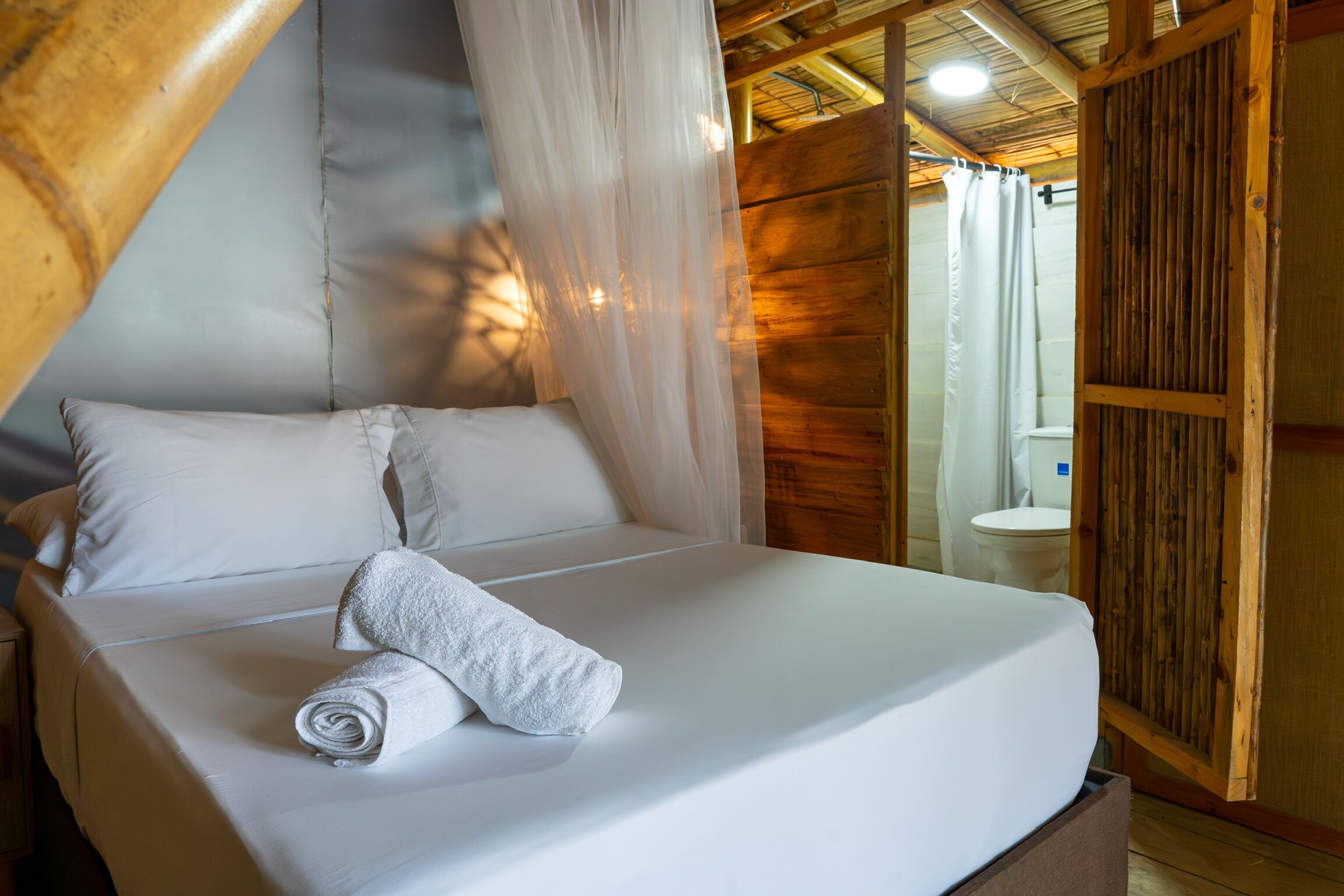 A bed with white linens and a rolled towel, a wooden ceiling, and a wooden wall.