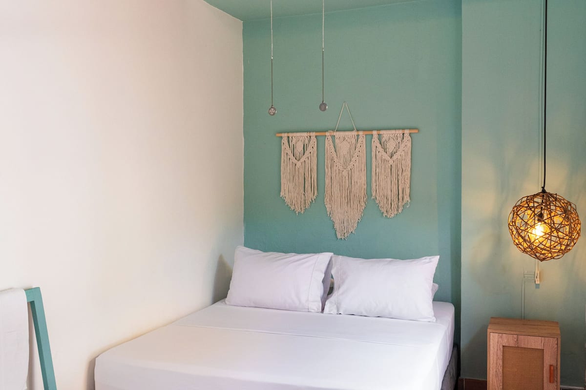 A bedroom with a bed, a wooden headboard, a macrame wall hanging, a hanging light fixture, and a small wooden cabinet.