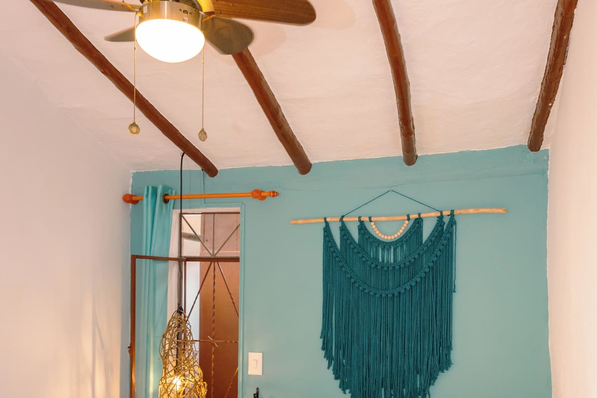 A bedroom with a bed, a ceiling fan, a macrame wall hanging, and a wooden nightstand.