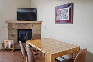 A living room with a fireplace, a flat-screen TV mounted above it, a framed picture on the wall, and a wooden dining table with chairs.