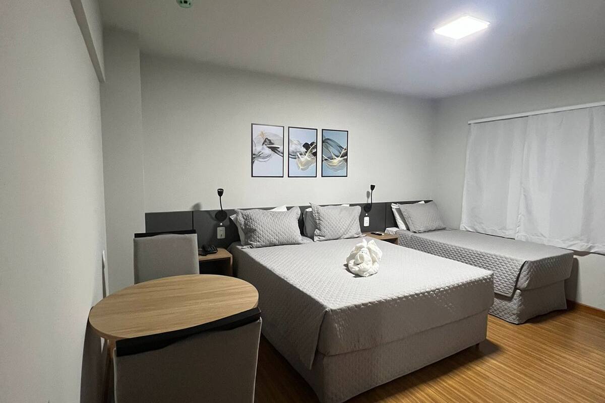 A modern hotel room with a large bed, a small desk, and a chair. There are three framed pictures on the wall and a window with white curtains.
