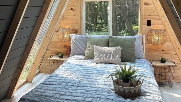 Une chambre confortable située sous les toits, avec un plafond en pente, un grand lit avec une literie à rayures, deux lampes de chevet et une fenêtre donnant sur la forêt.
