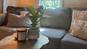 Un salon chaleureux avec un canapé gris, une petite table ronde agrémentée d’une plante et de bougies, et une fenêtre donnant sur un paysage verdoyant.