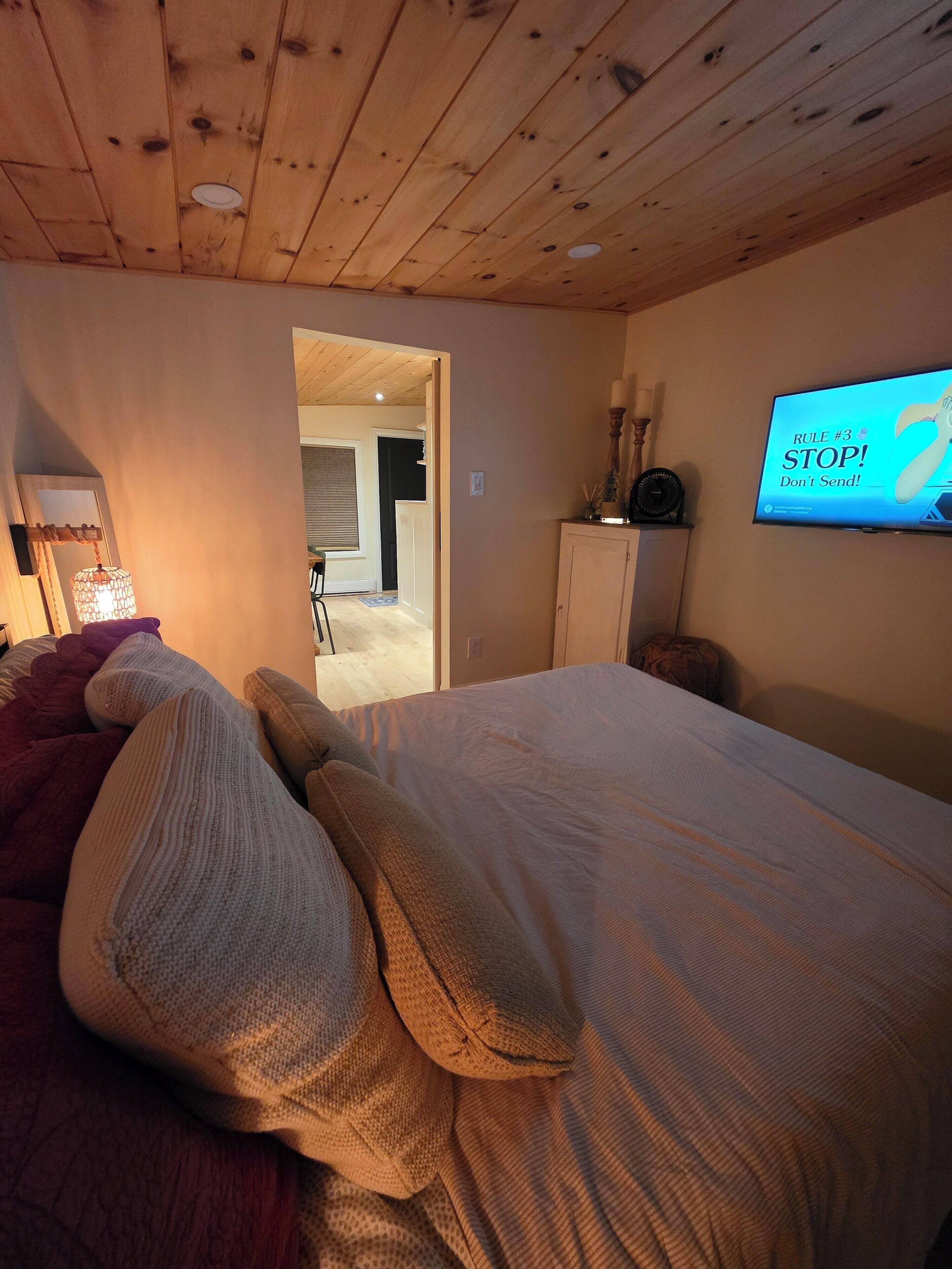 A bedroom with a wooden ceiling, a bed with pillows, a TV on the wall, and a door leading to another room.