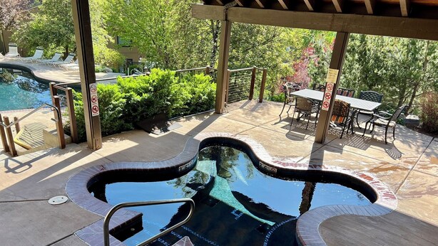 A hot tub with a unique design, surrounded by a covered patio area with outdoor furniture and a view of a pool and greenery. - Cozy Condo in Snowmass Village (Snowmass Village)