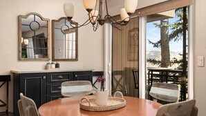 A dining area with a round table, chairs, a chandelier, and a large window with a view of trees. - Cozy Condo in Snowmass Village (Snowmass Village)