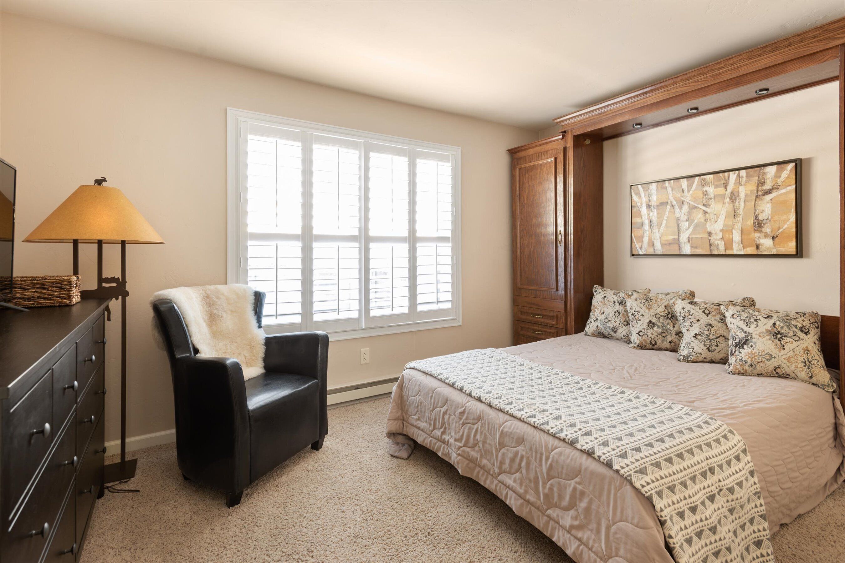 Une chambre avec une grande fenêtre, un lit agrémenté d’oreillers à motifs, un fauteuil recouvert d’un plaid en fausse fourrure, une commode et une lampe.