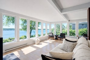 Une pièce spacieuse avec de grandes fenêtres donnant sur un lac et une végétation luxuriante. On y trouve un canapé blanc agrémenté d’un coussin décoratif texturé et une petite table d’appoint.