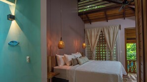 A bedroom with a bed, wooden headboard, hanging pendant lights, and a view of greenery outside. - Tucan Chiquita Beach Casitas (Playa Chiquita)